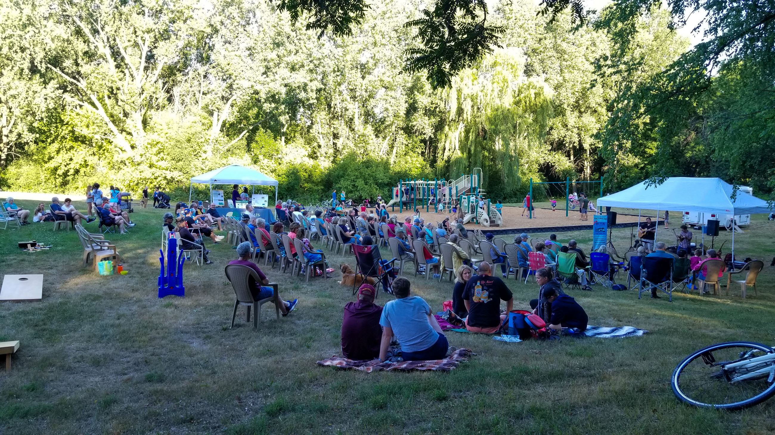 Crowd watches live music performance on a grassy hill with a playground in the background
