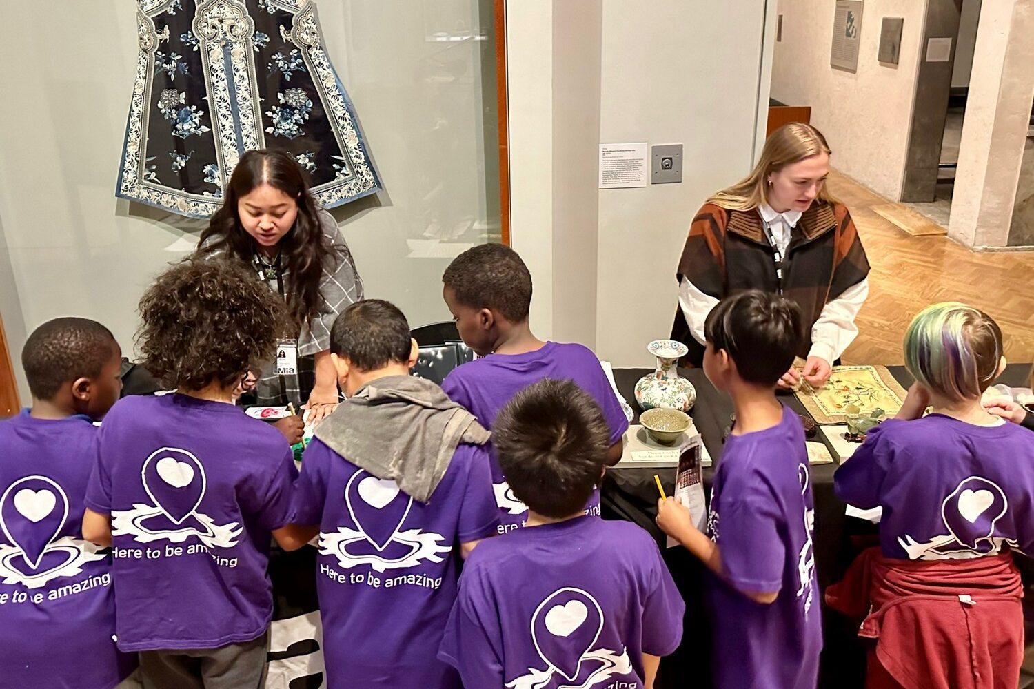 Students wearing purple T-shirts standing around tables with art objects. Two women are behind the tables talking to the students.